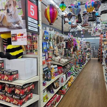 Pet store aisle with colorful pet toys and supplies. Hanging decorations include hot air balloons. A sale sign highlights a discount on an item.