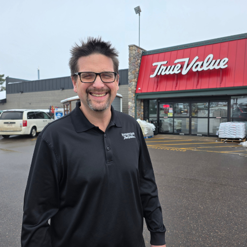 Man smiling broadly in front of a True Value hardware store. He wears glasses and a black shirt. The scene is bright, evoking a welcoming atmosphere.