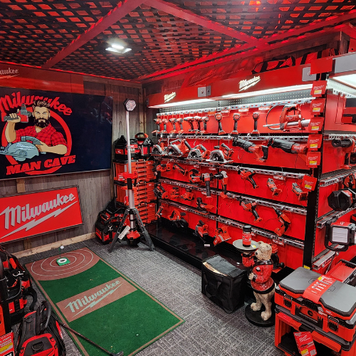 A well-organized man cave features an array of neatly displayed red Milwaukee power tools on shelves. The setting is enhanced by a branded sign and a cozy, focused ambiance.