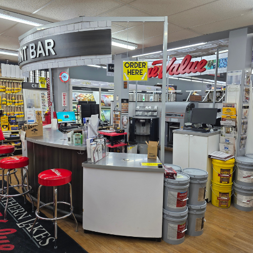 Interior of a hardware store featuring a pint bar with red stools, paint cans stacked nearby, and bright 'Order Here' signs. The atmosphere is organized and bustling.