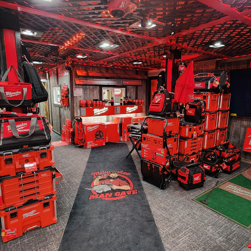 A well-organized man cave features an array of neatly displayed red Milwaukee power tools on shelves. The setting is enhanced by a branded sign and a cozy, focused ambiance.