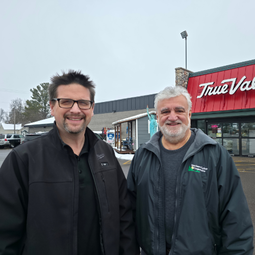 Two men stand smiling in front of a True Value store on a cloudy day. They are dressed in dark jackets, conveying a friendly and casual atmosphere.