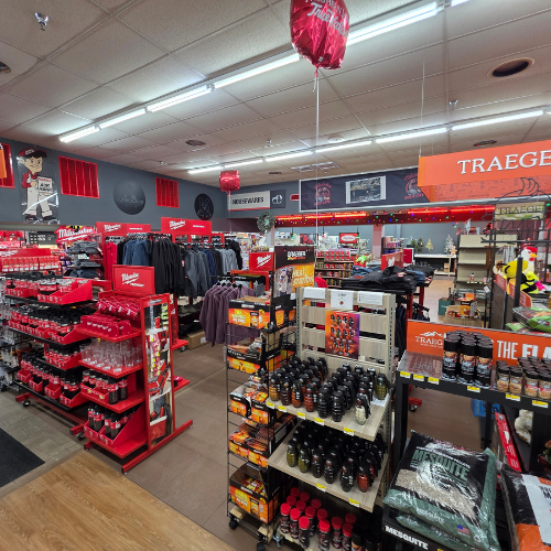 A hardware store interior with shelves of tools, clothing, and grilling supplies. Bright red displays are prominent, creating a bustling, organized feel.