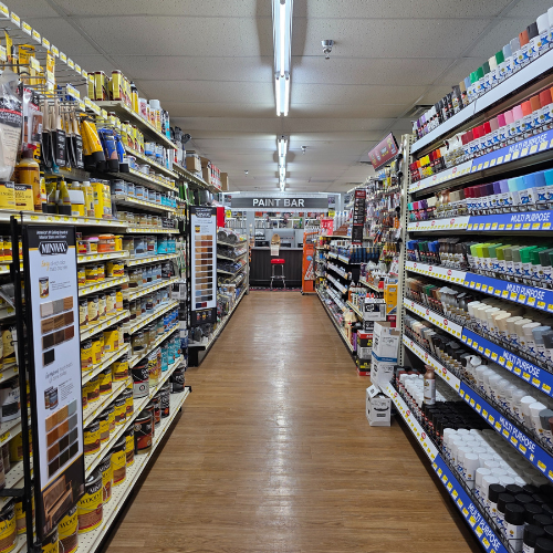 A well-organized hardware store paint aisle with shelves full of colorful paint cans, brushes, and supplies. The bright lighting creates a welcoming atmosphere.