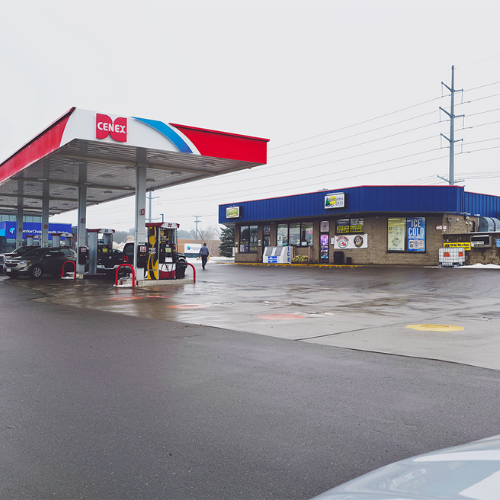 A quiet gas station with a red and white canopy on an overcast day. A few cars are parked, and a convenience store with a blue roof is visible.