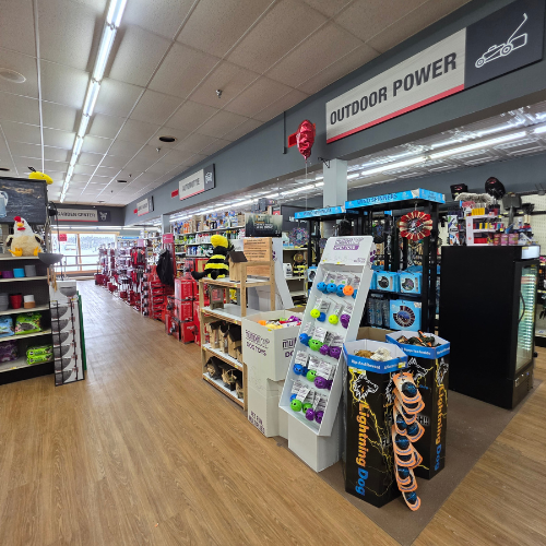 A well-lit store aisle features power tools and lawn equipment. Shelves are neatly stocked with colorful products. Signs read 