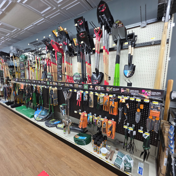 A hardware store aisle with neatly arranged gardening tools on display. Various shovels, rakes, trowels, and accessories create an organized, colorful scene.
