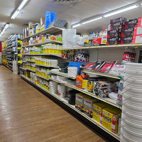 A hardware store aisle with wooden flooring filled with cleaning supplies, buckets, and gloves. The shelves are neatly stocked, creating a busy and organized atmosphere.