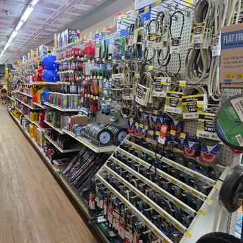Aisle in a hardware store lined with various tools and supplies on shelves, including cables, tapes, and oil cans. Bright lighting, organized display.