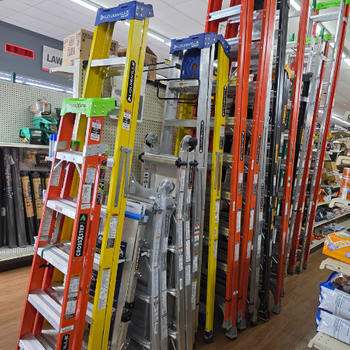 A variety of ladders in orange, yellow, and silver are displayed upright in a hardware store aisle, creating a colorful and organized scene.