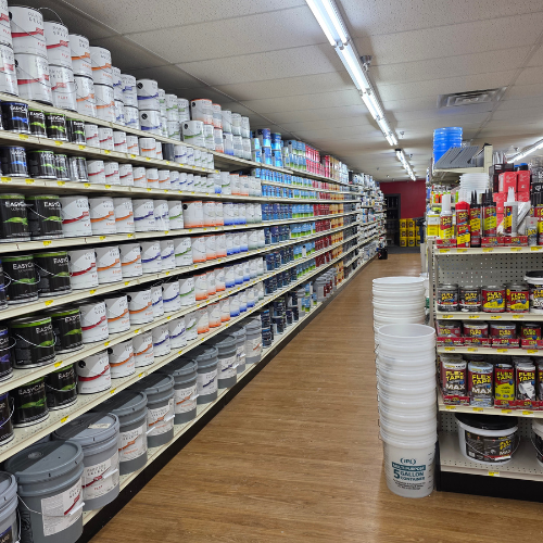 A hardware store aisle filled with stacked paint cans on the left and various supplies on the right. Fluorescent lights illuminate the wooden floor.