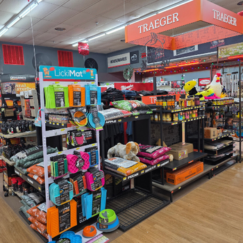 Pet store aisle with colorful dog toys, treats on a LickiMat display. Nearby, pet food bags and Traeger grill accessories under warm lighting.