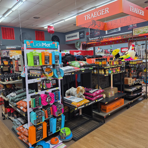 Pet store aisle with colorful dog toys, treats on a LickiMat display. Nearby, pet food bags and Traeger grill accessories under warm lighting.