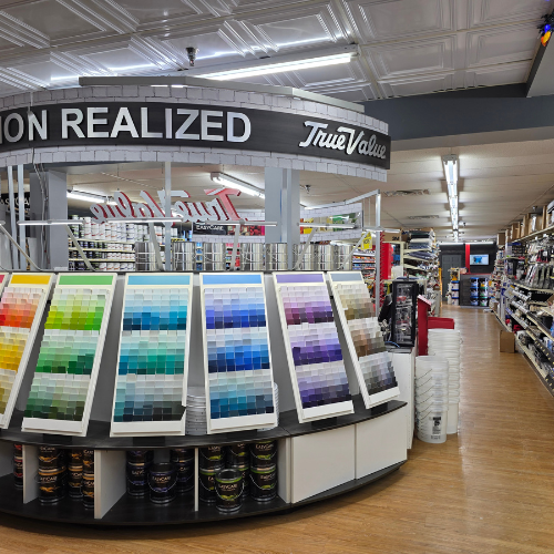 A vibrant display of paint sample cards arranged by color on a circular stand in a hardware store aisle. Overhead signage reads 